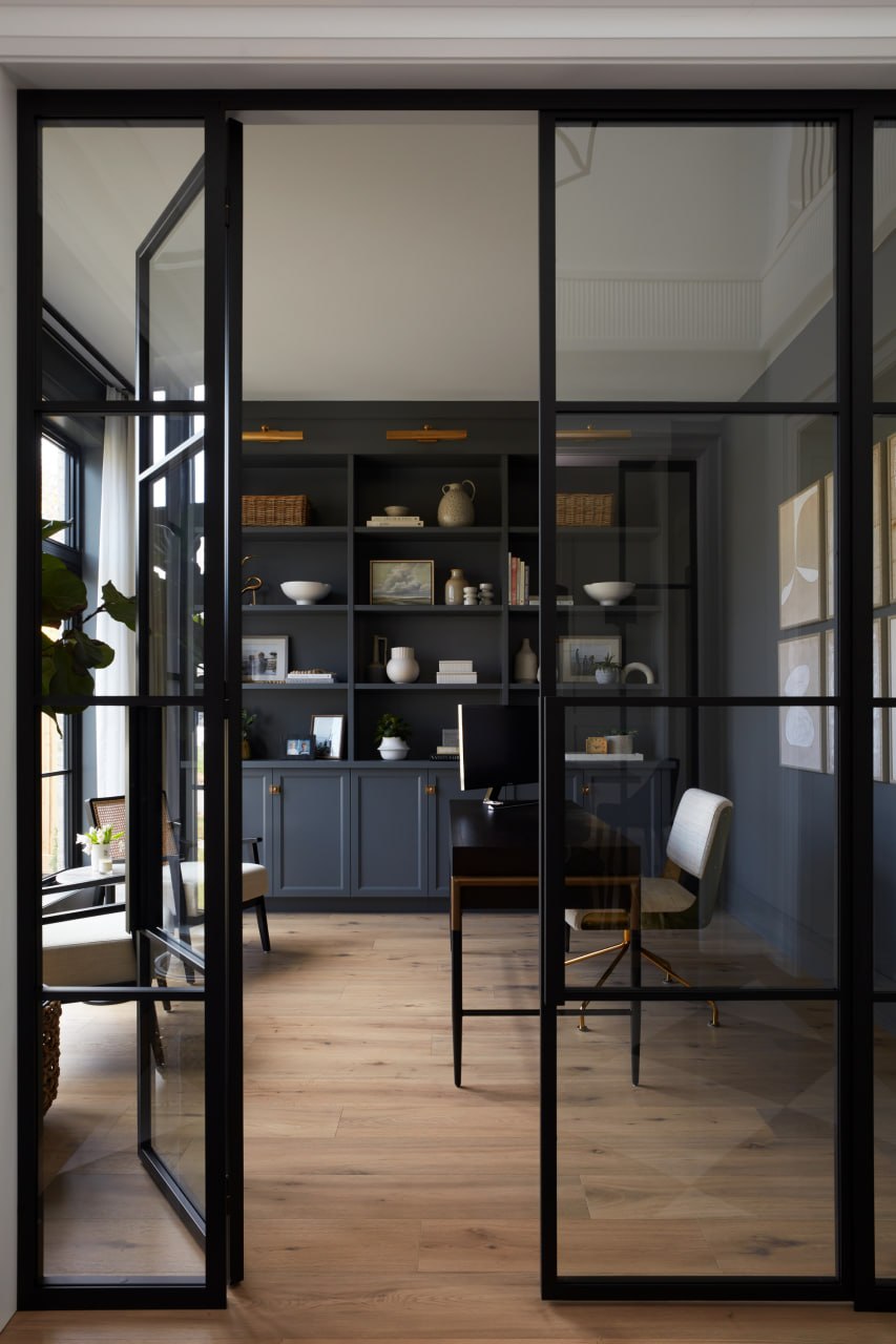 Custom home office with navy built-in shelving, brass picture lights and steel-and-glass doors