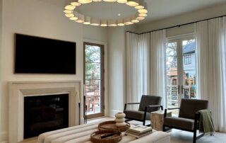 Warm minimalist living room in luxury Oakville custom home featuring neutral palette, modern fireplace, sculptural ottoman, circular chandelier, and floor-to-ceiling drapery by Prasada Custom Homes.